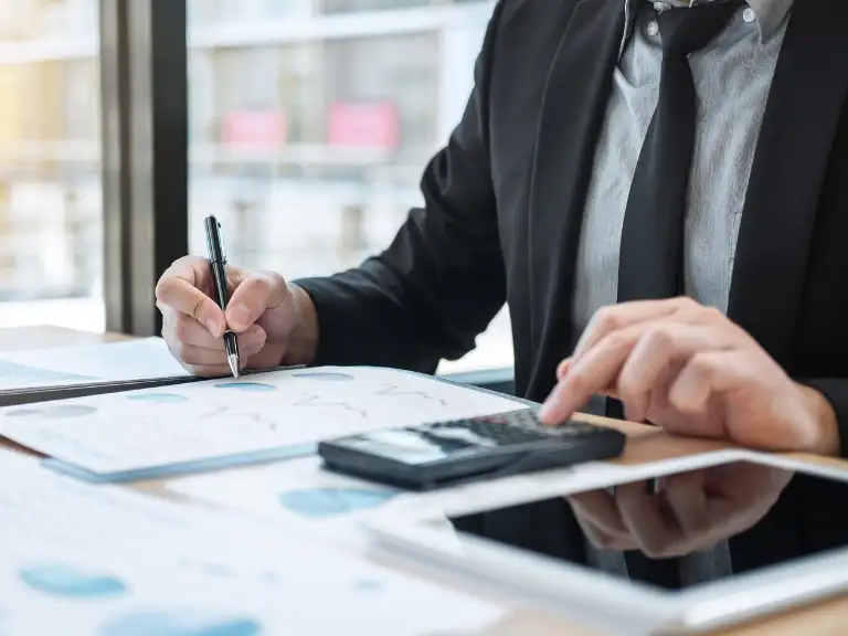 Um profissional de terno e gravata utilizando uma calculadora e escrevendo em documentos com gráficos, com um tablet ao lado